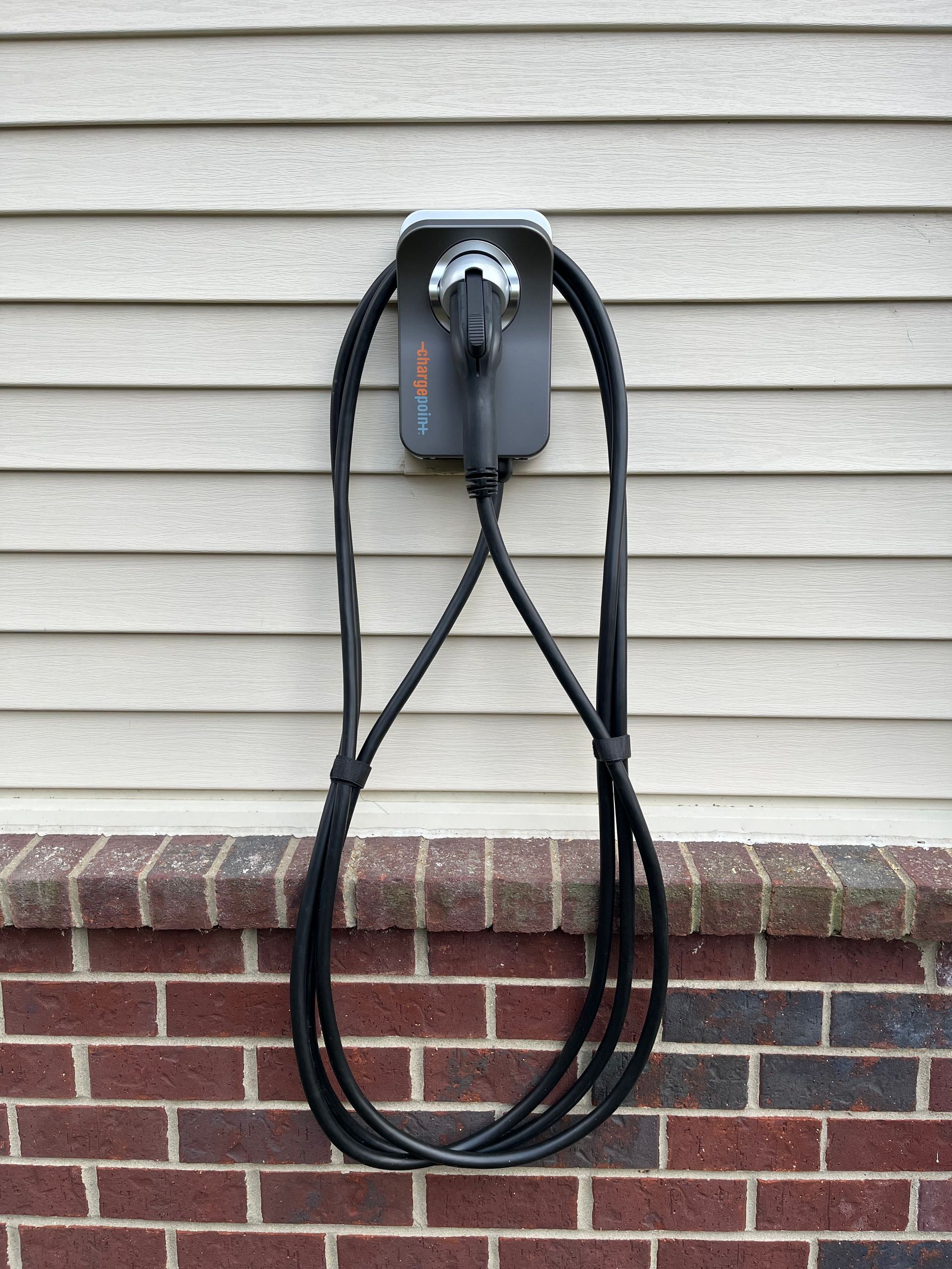 Wall-mounted electric vehicle charger with black cable coiled on beige siding and brick wall