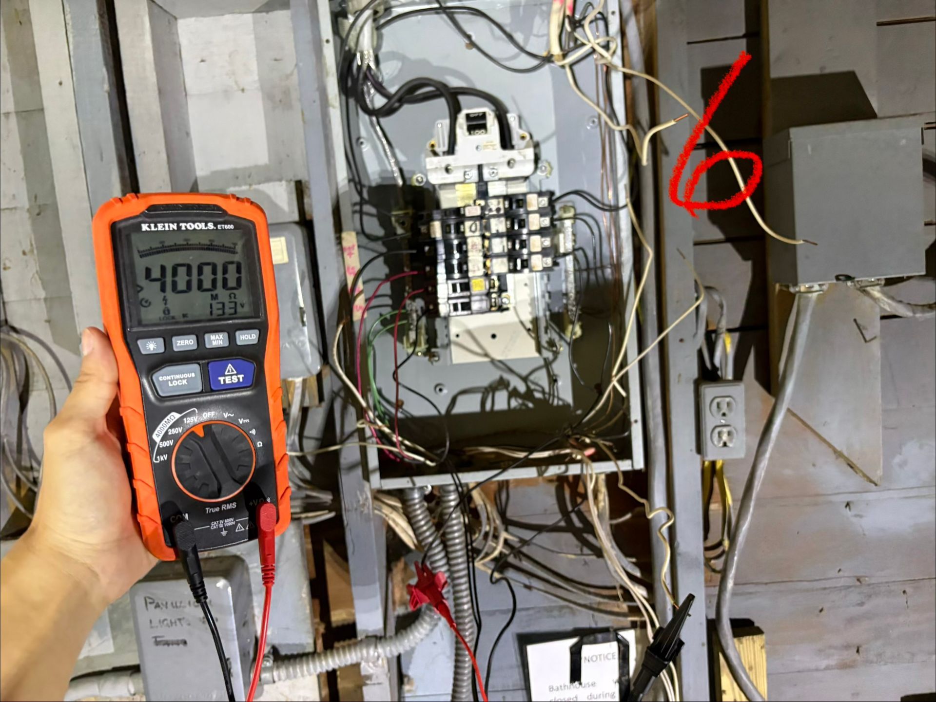 Handheld multimeter testing wiring inside an electrical panel, with a red arrow marking a wire on the right.