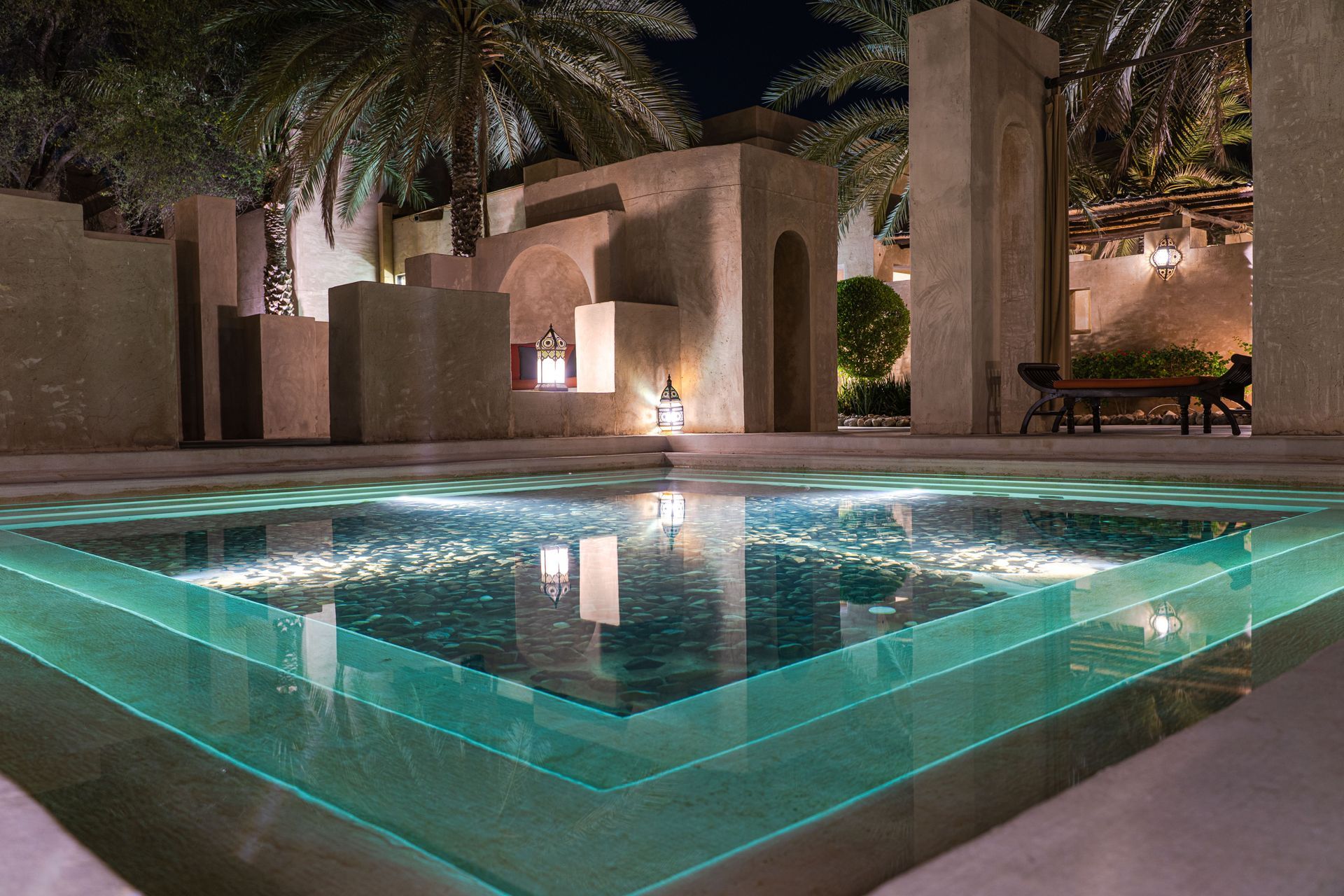Illuminated modern courtyard with a turquoise-lit reflecting pool and palm trees at night
