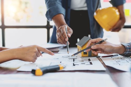 Hands drafting a floor plan with tools and hard hats on a table