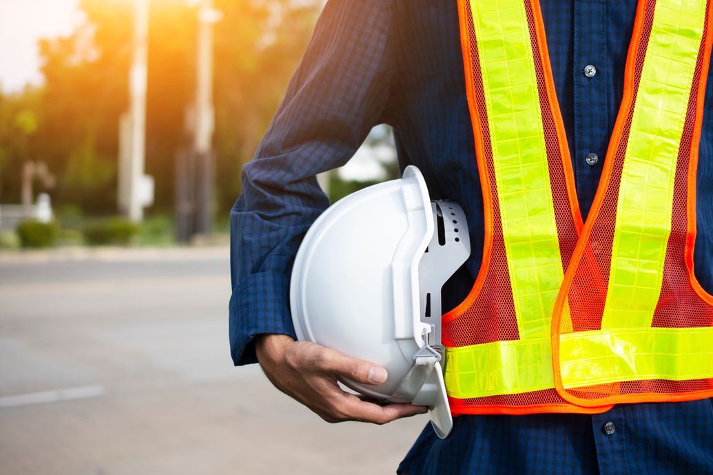 Construction worker in reflective vest holding a white hard hat outdoors at a work site