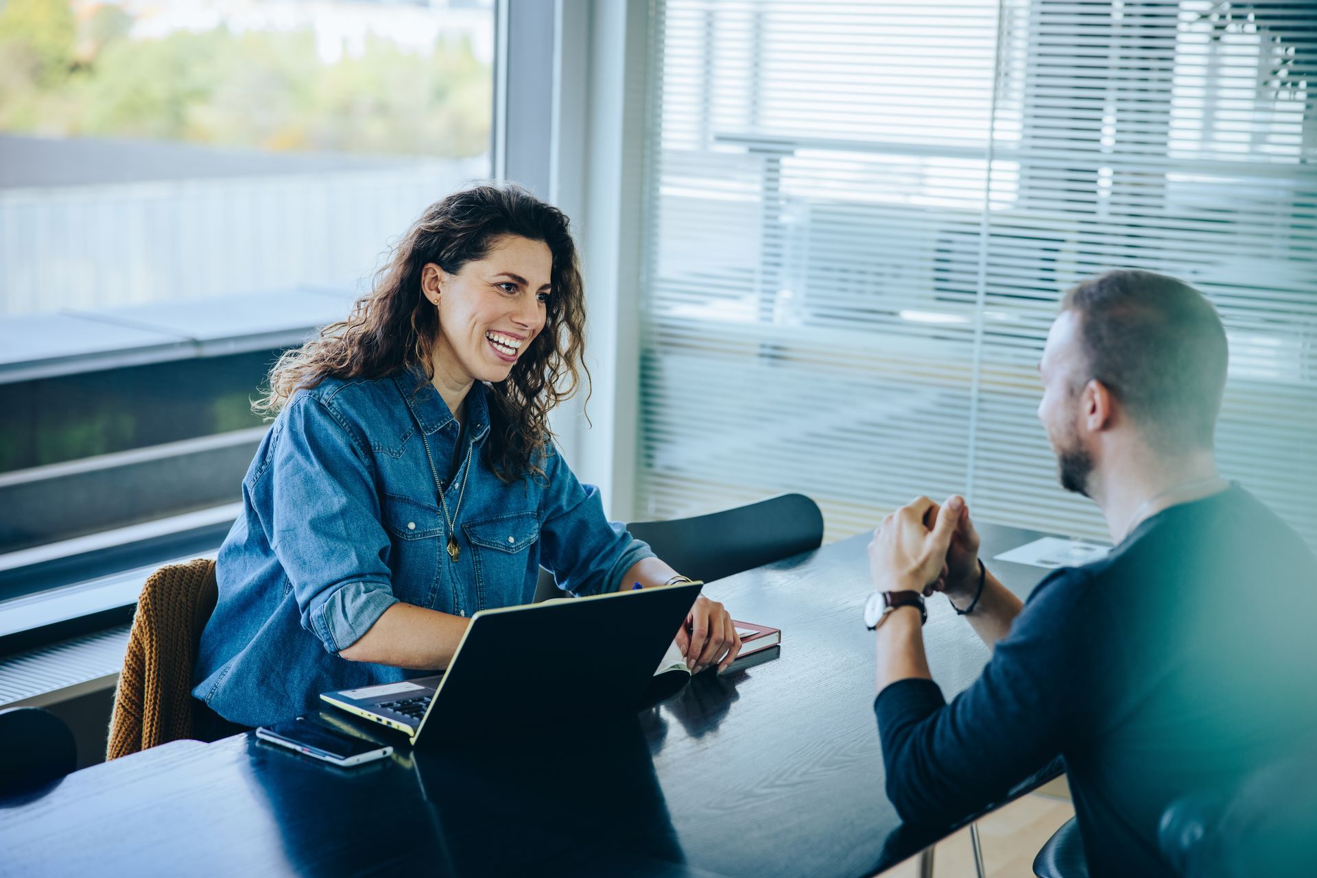 Two people talking across a conference table in a bright office, with a laptop and microphone on the table.