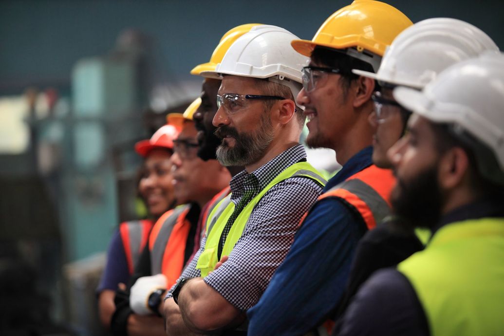 Workers in hard hats and safety vests standing in a factory, listening attentively.