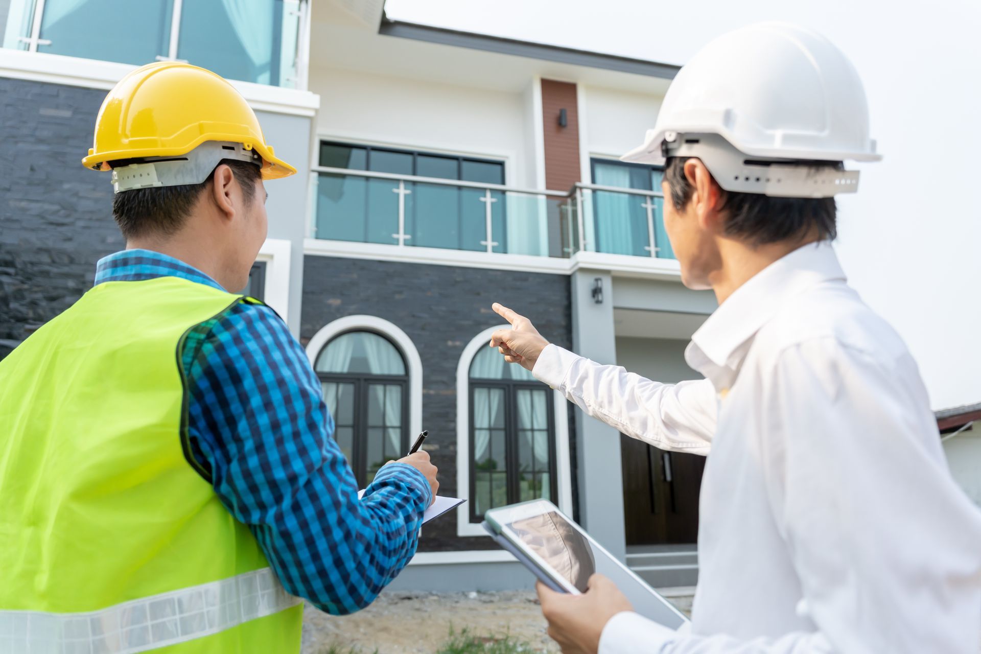 Two construction workers in hard hats inspect a house exterior with a tablet and gesture toward the building.