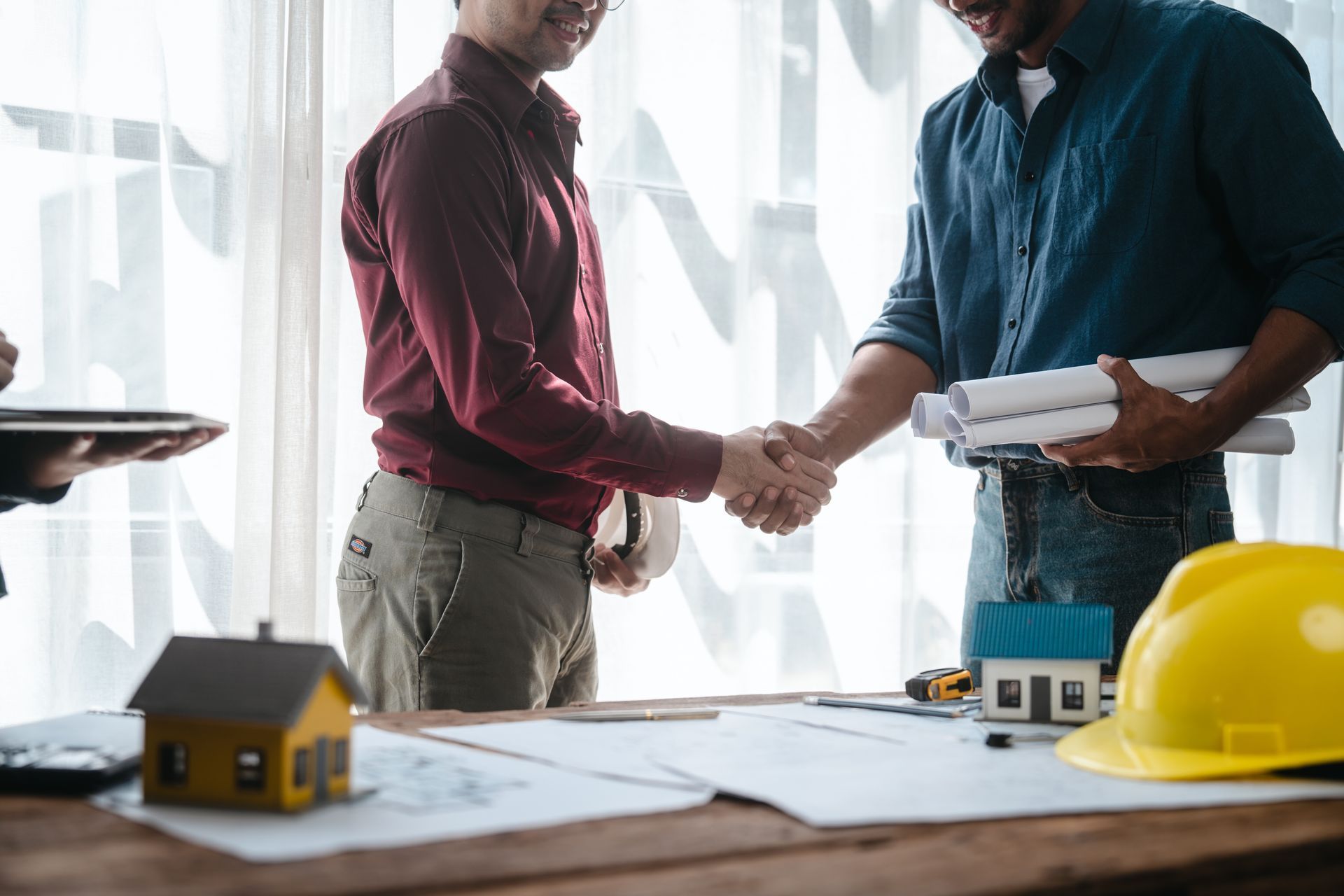 Two people shaking hands over architectural plans with a yellow hard hat on the table
