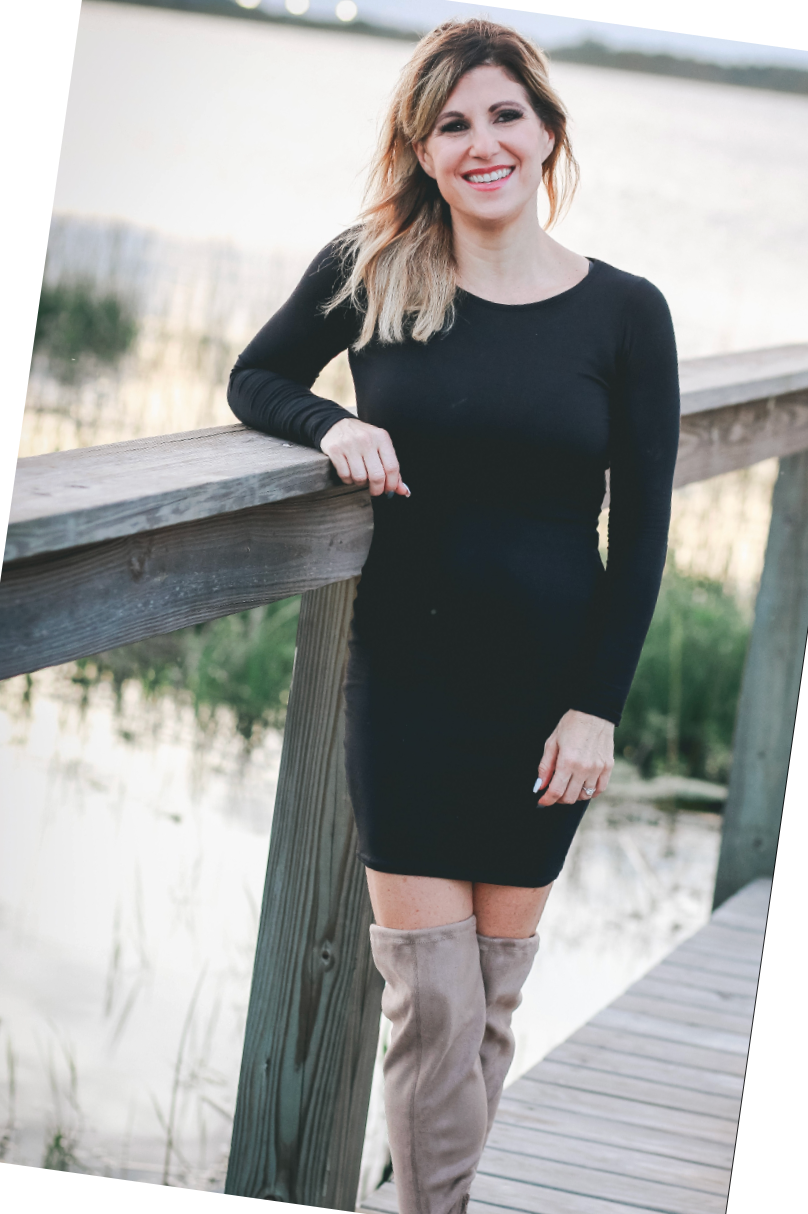 Woman in a black dress leaning on a wooden bridge railing outdoors, smiling at the camera.