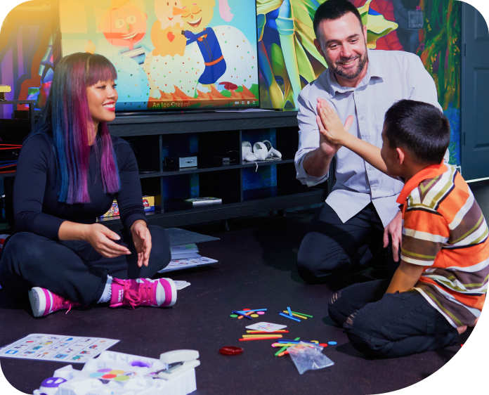 Teacher and two children sitting on the floor, laughing and playing with colorful cards in a classroom
