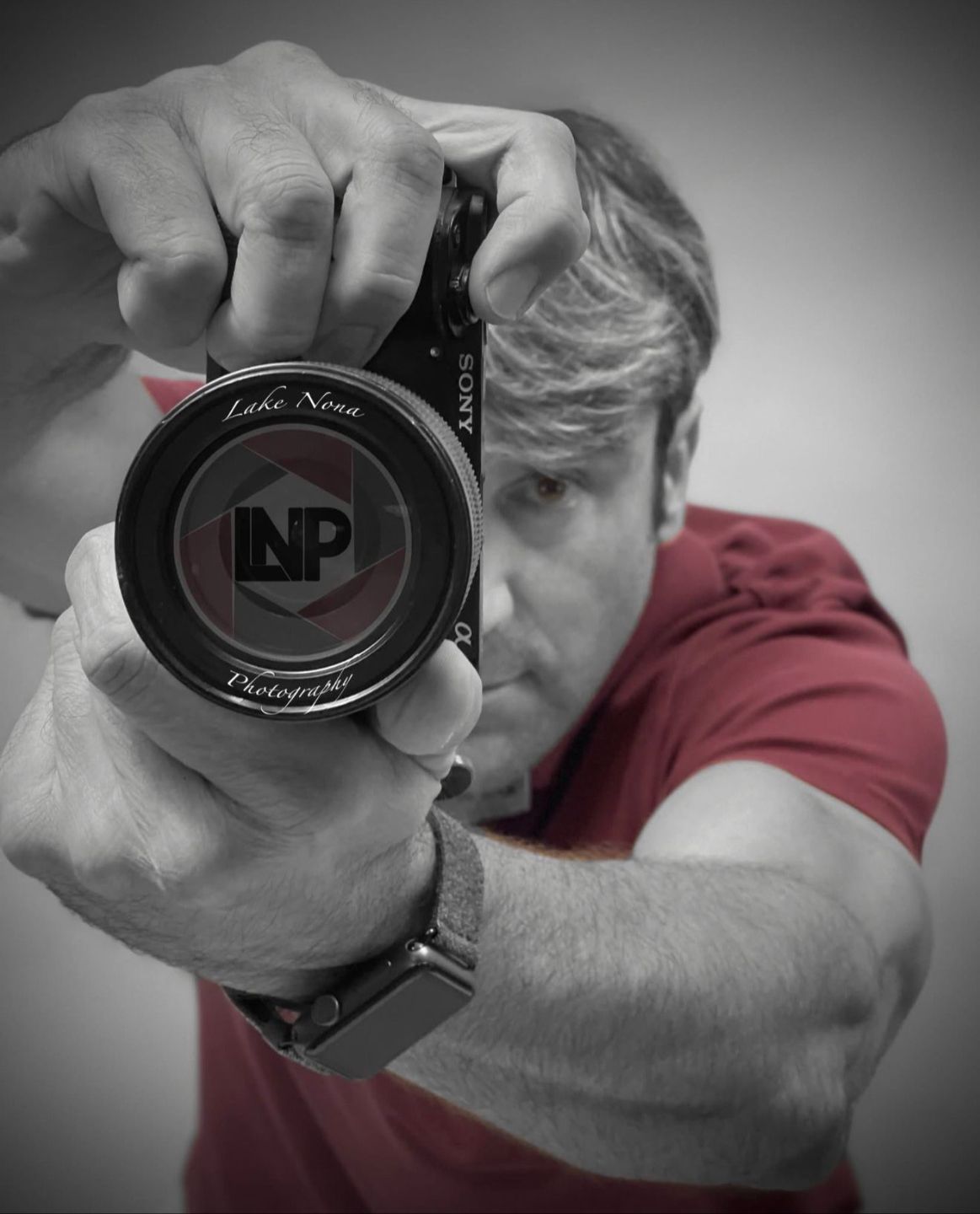 Person aiming a camera at the viewer, with a red shirt and black-and-white background.