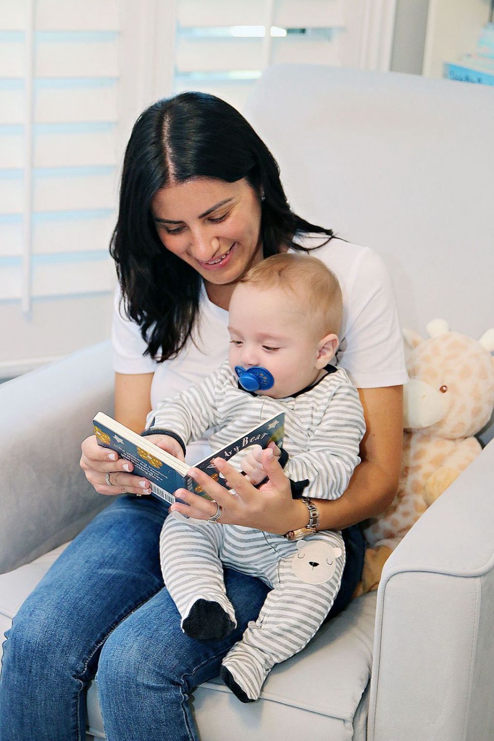 Woman reading a book to a baby on a white chair in a bright room