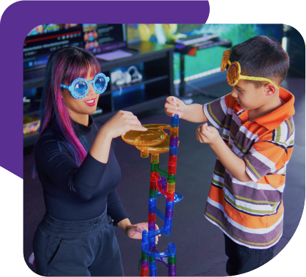 Two children wearing colorful goggles play with a tall plastic marble run indoors.