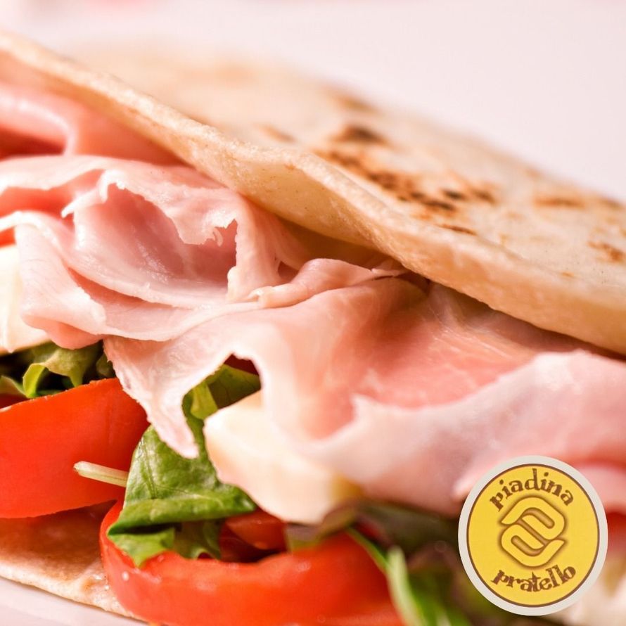 Primo piano di un panino con prosciutto, pomodoro e lattuga su pane piatto con una piccola etichetta con il logo.