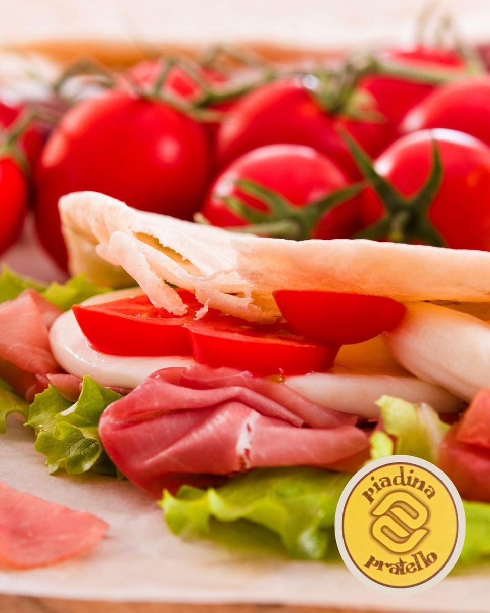 Tramezzino aperto con pomodoro, lattuga e affettati davanti a pomodorini ciliegino.