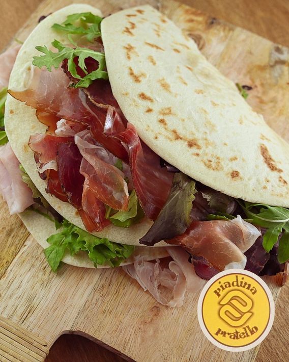 Panino con focaccia, prosciutto e lattuga, servito su un tagliere di legno, con un cartellino rotondo del menù.