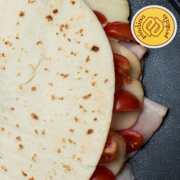 Primo piano di un panino con pane piatto, pomodori, prosciutto e formaggio, sormontato da una piccola etichetta gialla.