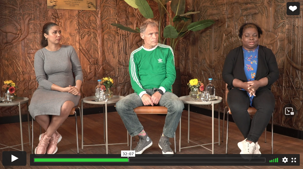 Drie mensen zitten voor een gesprek. Op de achtergrond is een houten lambrisering te zien met planten en bloemen.