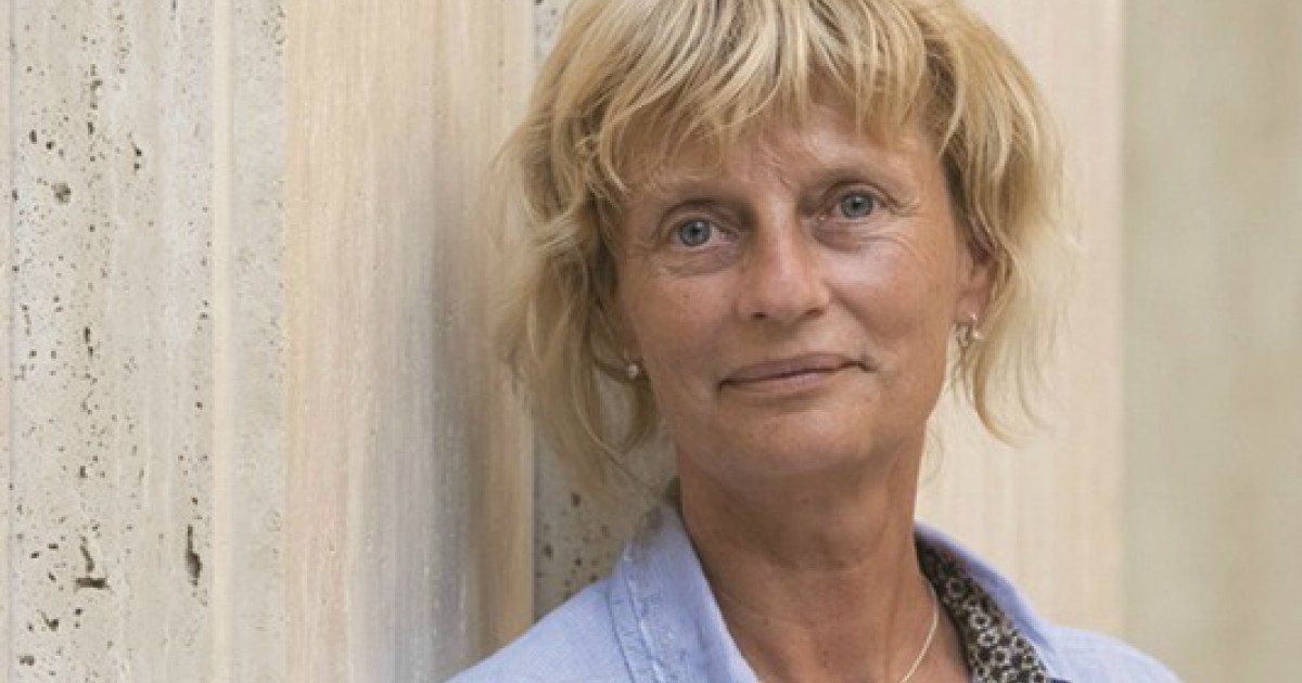 Een vrouw met kort blond haar en een blauwe blazer leunt tegen een muur met structuur en glimlacht lichtjes.