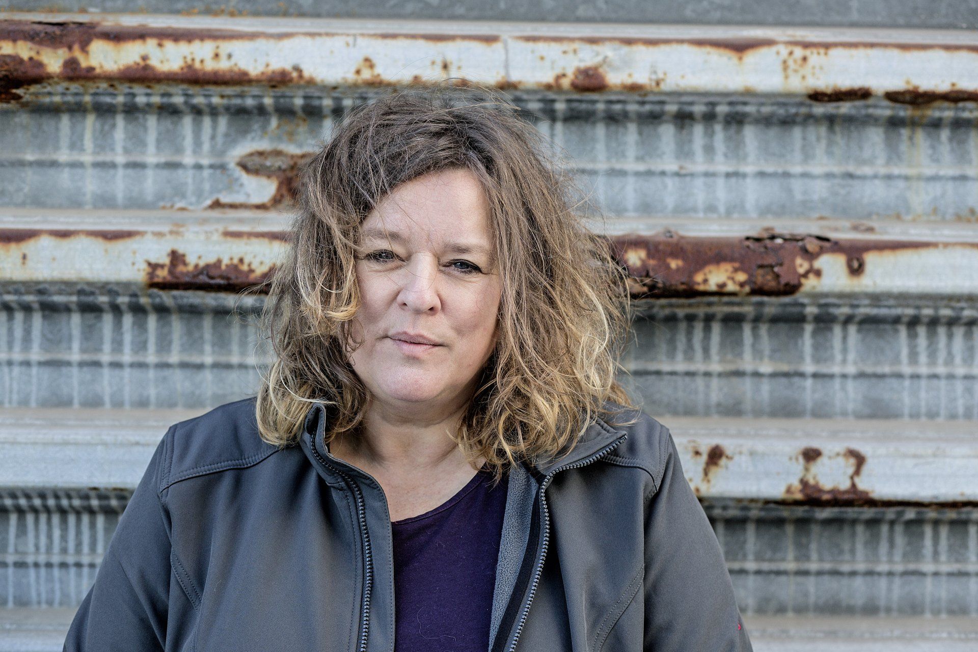 Vrouw met warrig bruin haar en een grijze jas tegen een roestige, gegolfde metalen achtergrond.