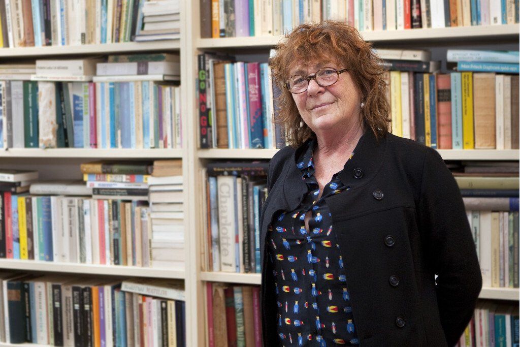 Vrouw staat voor een boekenplank, gekleed in een zwarte jas en een blouse met patroon.