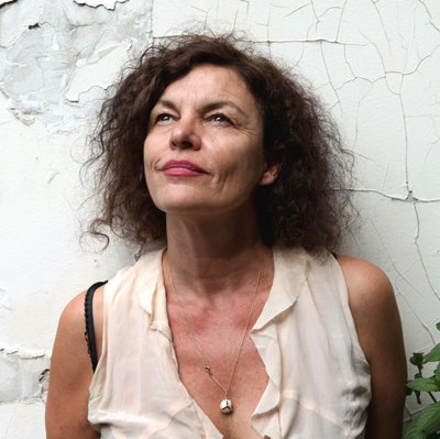 Een vrouw met krullend bruin haar kijkt omhoog, gekleed in een licht topje en met een ketting om, tegen een afbladderende witte muur.