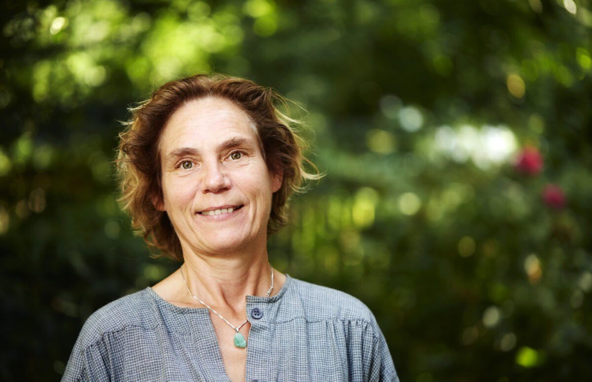 Een vrouw met bruin haar, gekleed in een blauwe blouse en met een ketting om, staat lachend buiten met groen gebladerte op de achtergrond.