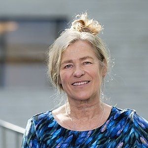 Vrouw met lichtbruin haar in een knotje, lachend, gekleed in een blauw topje met patroon, buiten.