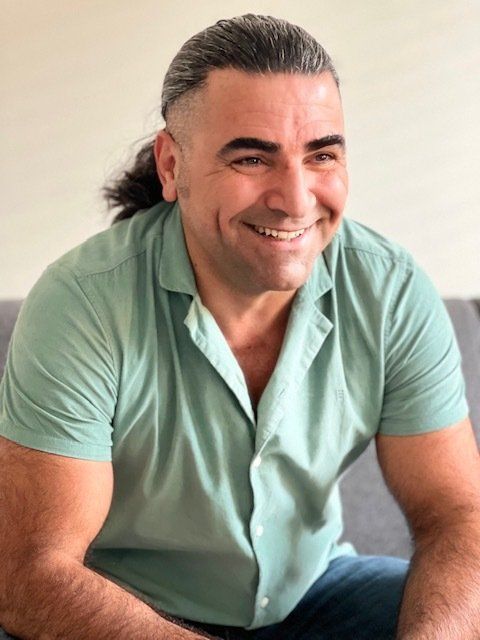 Een man met donker haar dat naar achteren is gebonden, glimlacht, draagt een groen shirt en zit binnen.