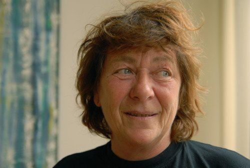 Een vrouw met kort, warrig bruin haar glimlacht en kijkt niet in beeld. Ze staat voor een schilderij met blauwe en groene tinten.