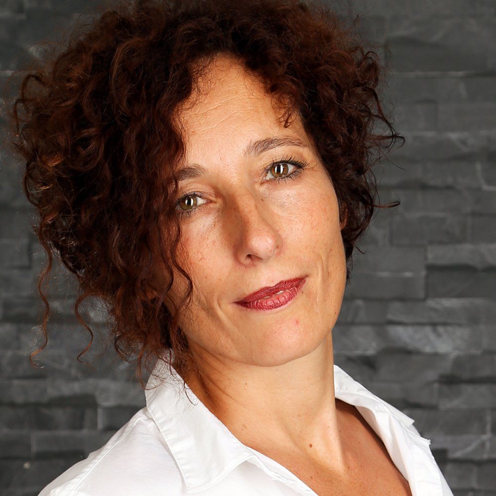Vrouw met krullend bruin haar, gekleed in een witte blouse, glimlacht lichtjes tegen een grijze muur.