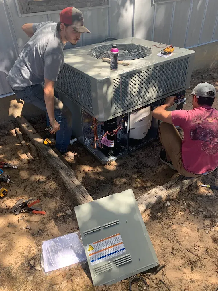 Two workers installing a large outdoor HVAC unit on a dirt slope, with a detached metal cover nearby.