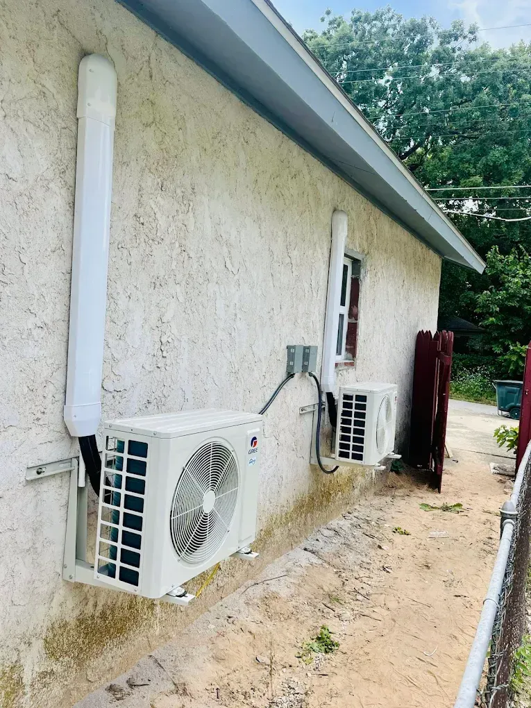 Two white outdoor AC units mounted on a stucco house wall beside a narrow dirt path.