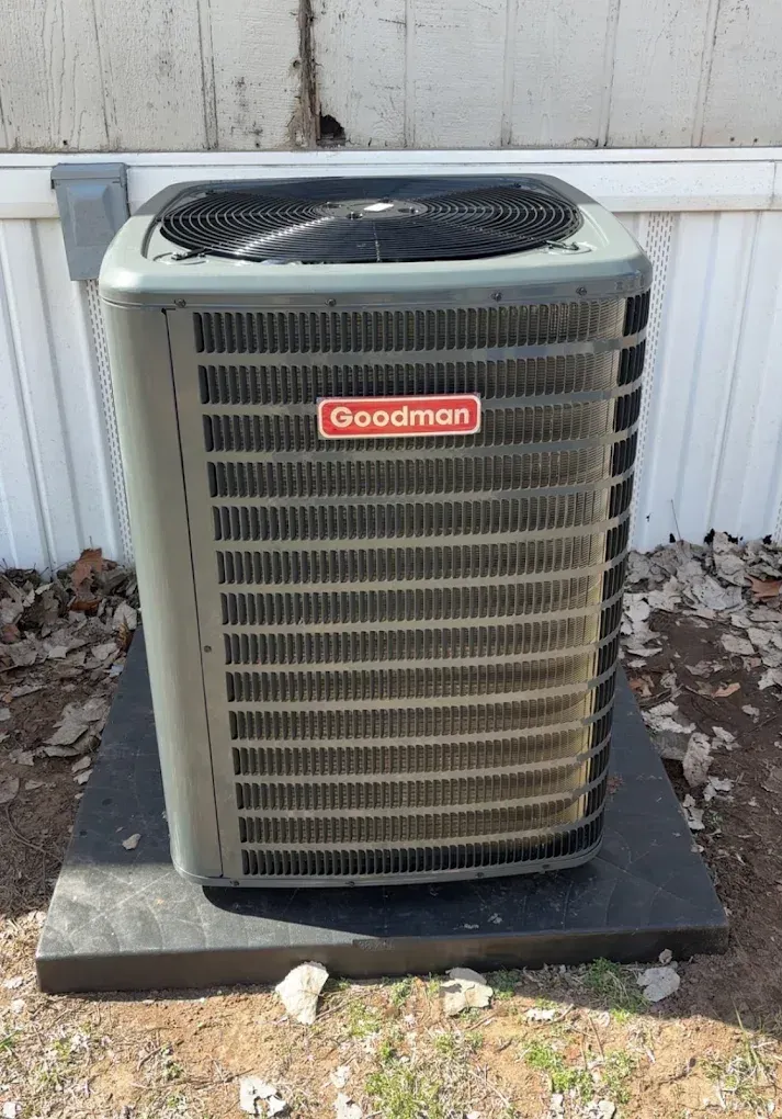 Outdoor Goodman air conditioner unit on a concrete pad beside a white fence and scattered leaves.