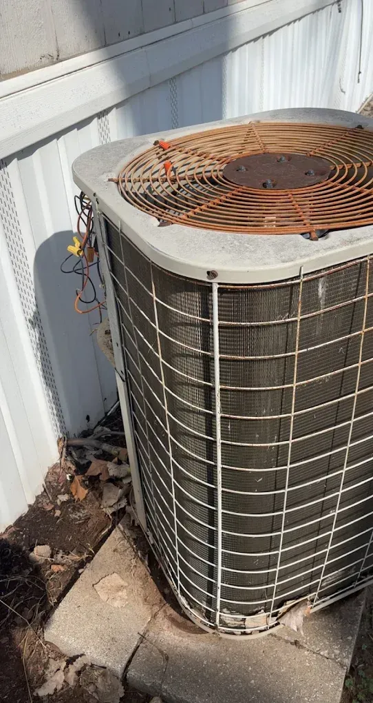 Outdoor air conditioner unit beside a white wall, with rust on the top grille and dirt around the base