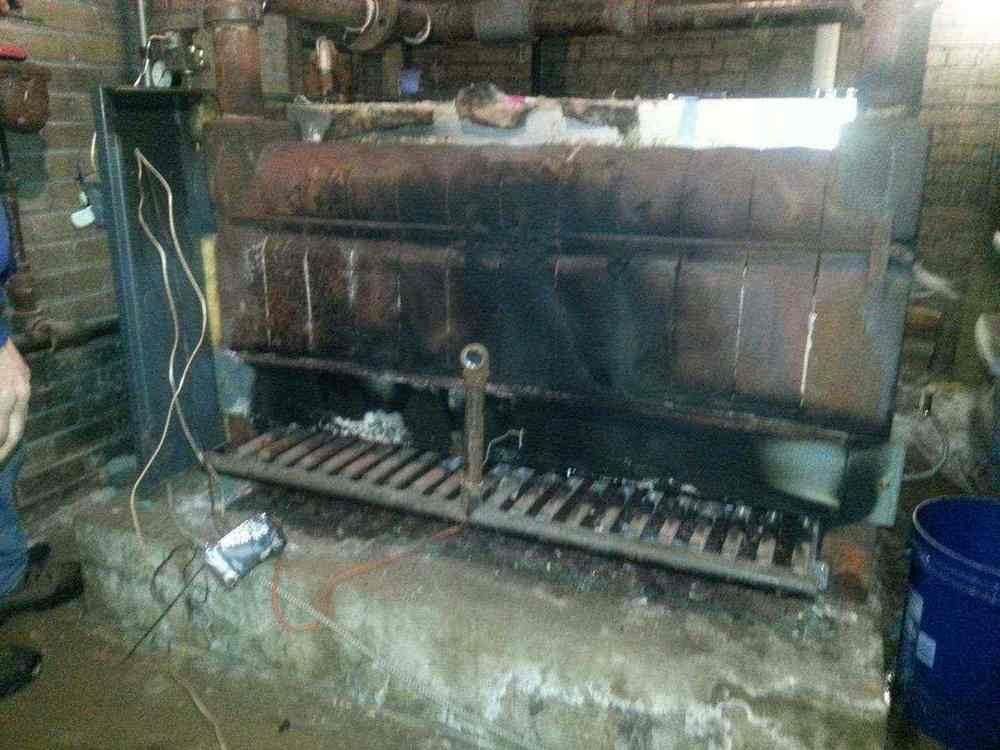 Coal-fired furnace in a brick-walled room. Black metal, ash-covered grate, water bucket at right.
