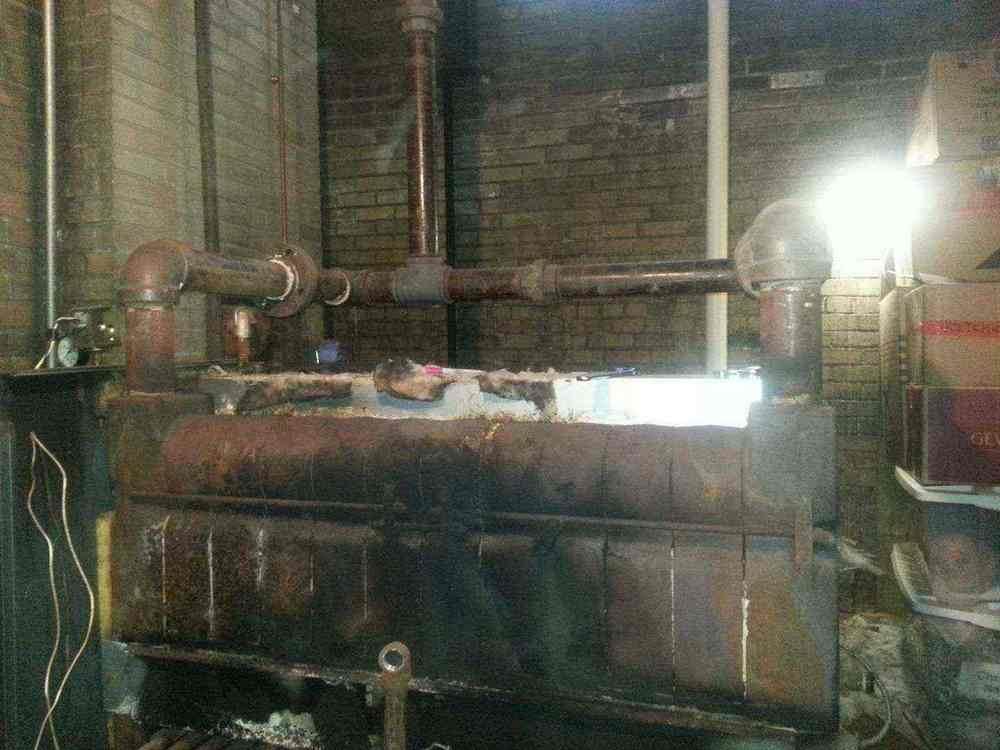 Rusty, old industrial boiler with pipes, set against brick wall in a dimly lit space.
