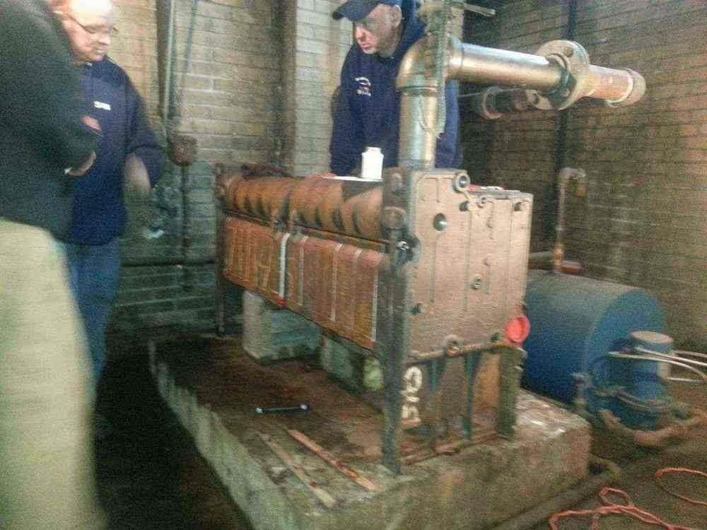 Two men examine a large, rusty industrial machine in a brick-walled room.