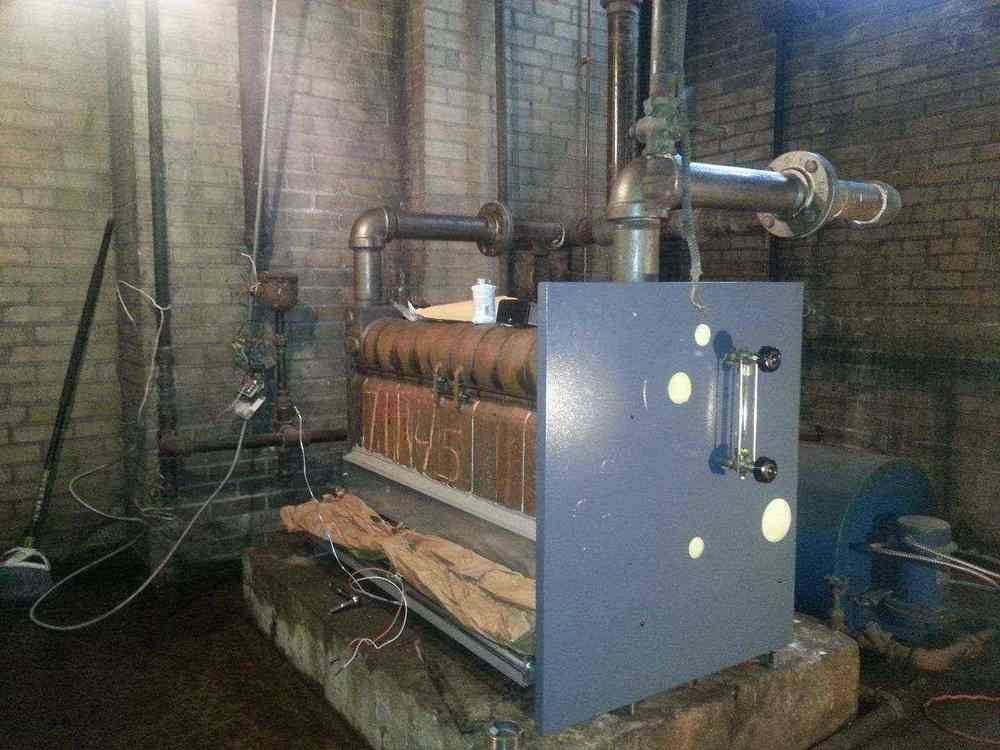 An industrial boiler with copper coils and metal pipes in a brick-walled basement.