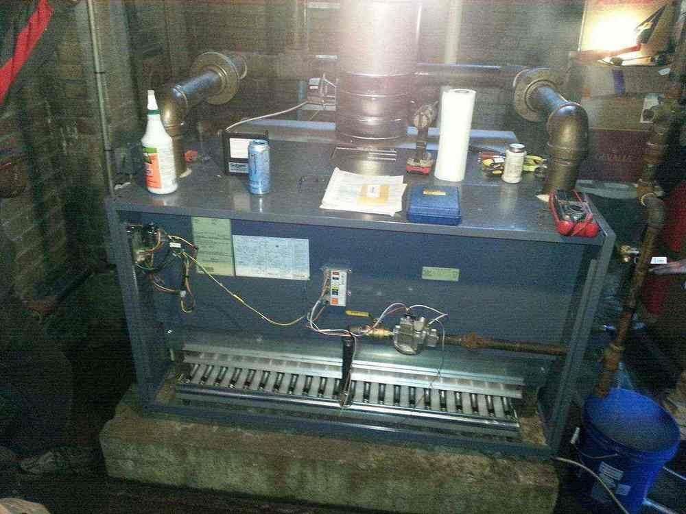 Blue furnace with exposed front, pipes, and tools on top, in a basement.