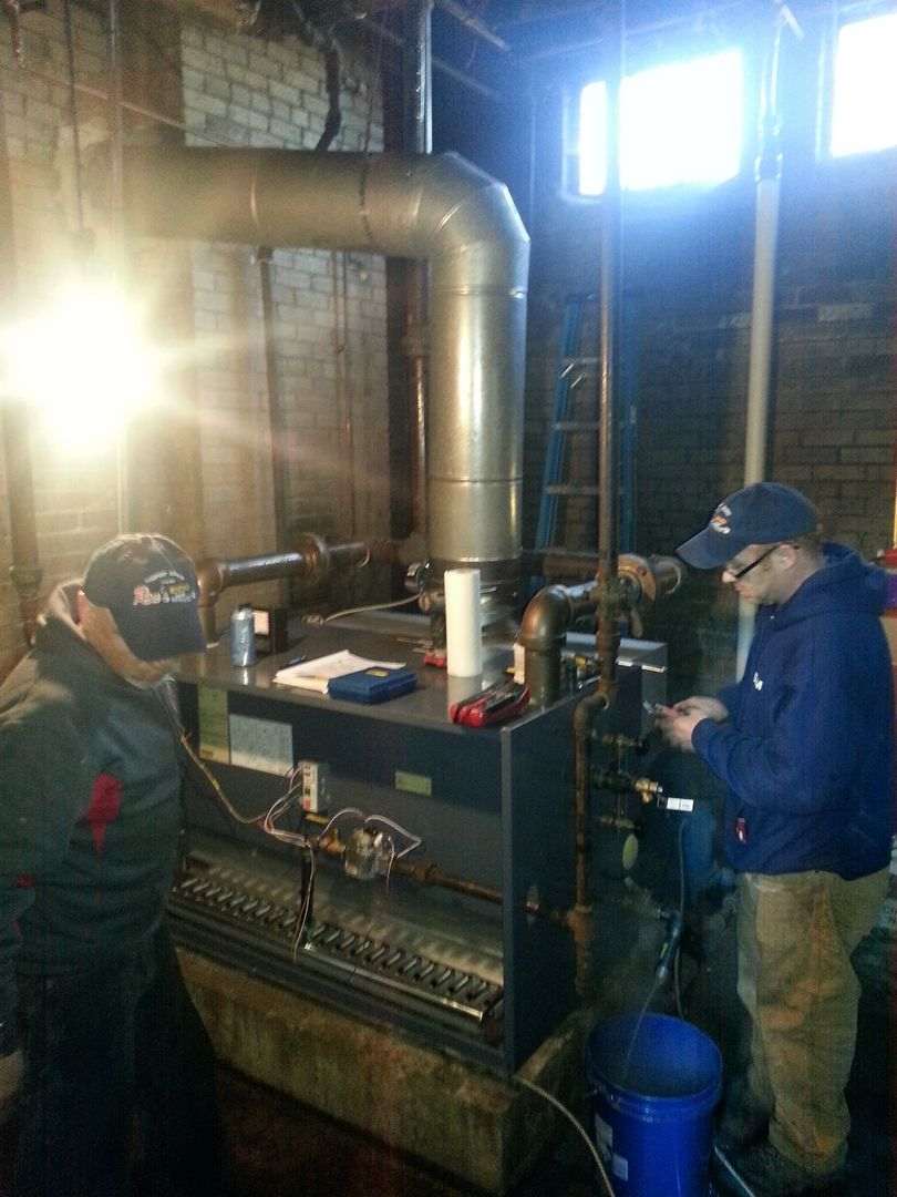 Two people working on a furnace in a basement. One adjusts a valve, the other observes. Gray, metal, and pipes.