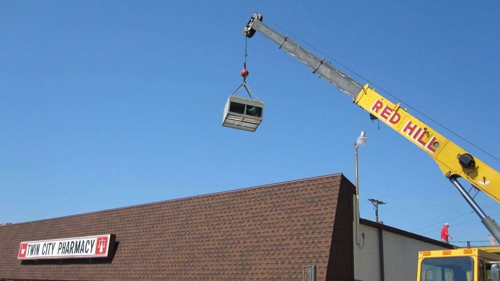 Crane lifting an HVAC unit above a brick building with a pharmacy sign against a blue sky.