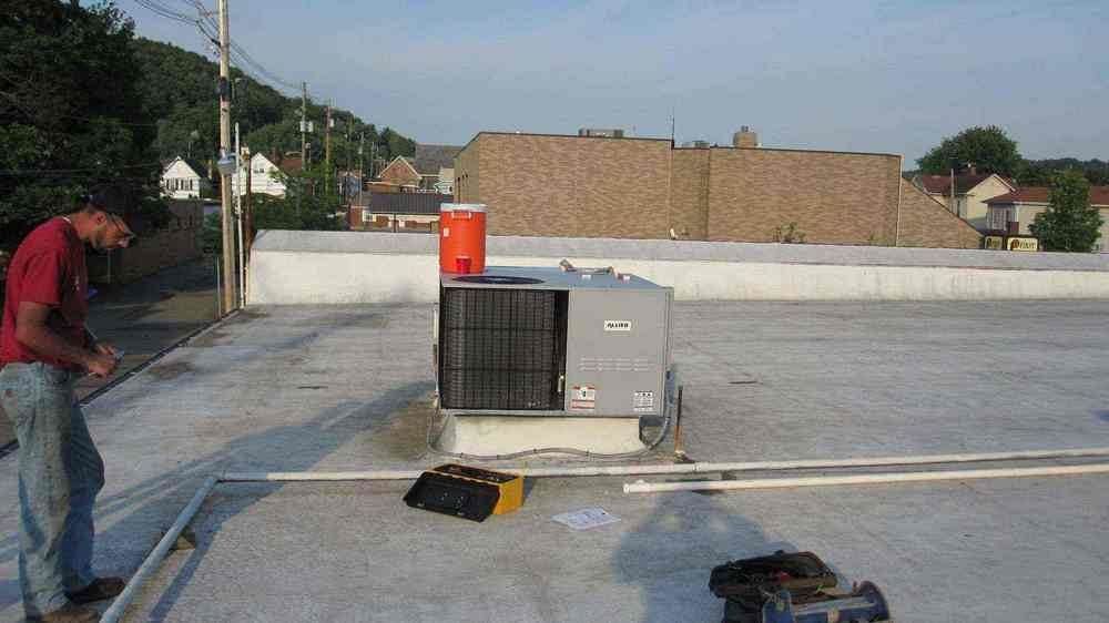 Man working on rooftop HVAC unit near a building and hillside; sunny day.