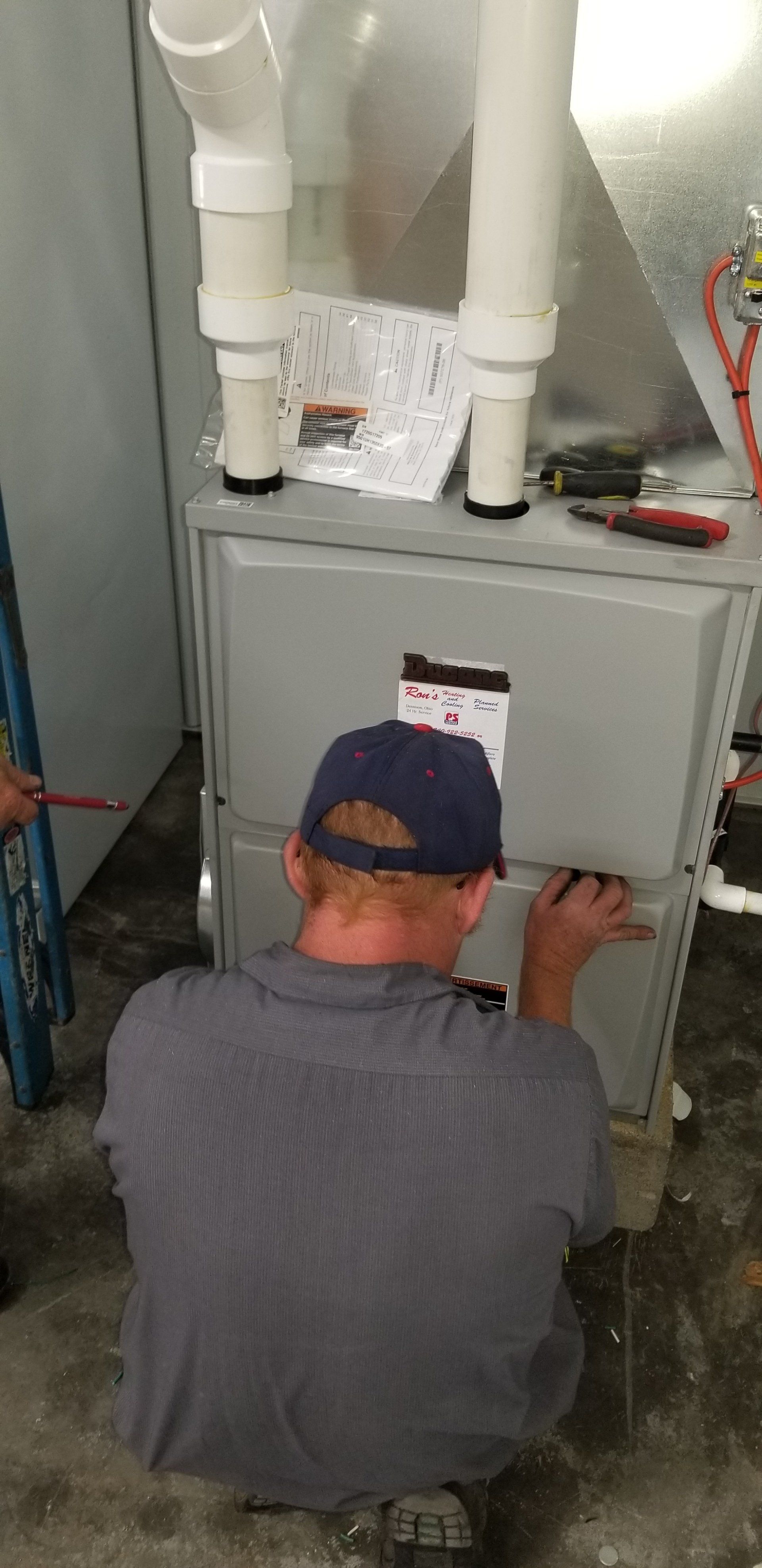 A person in a blue hat works on a furnace. White pipes are connected above.