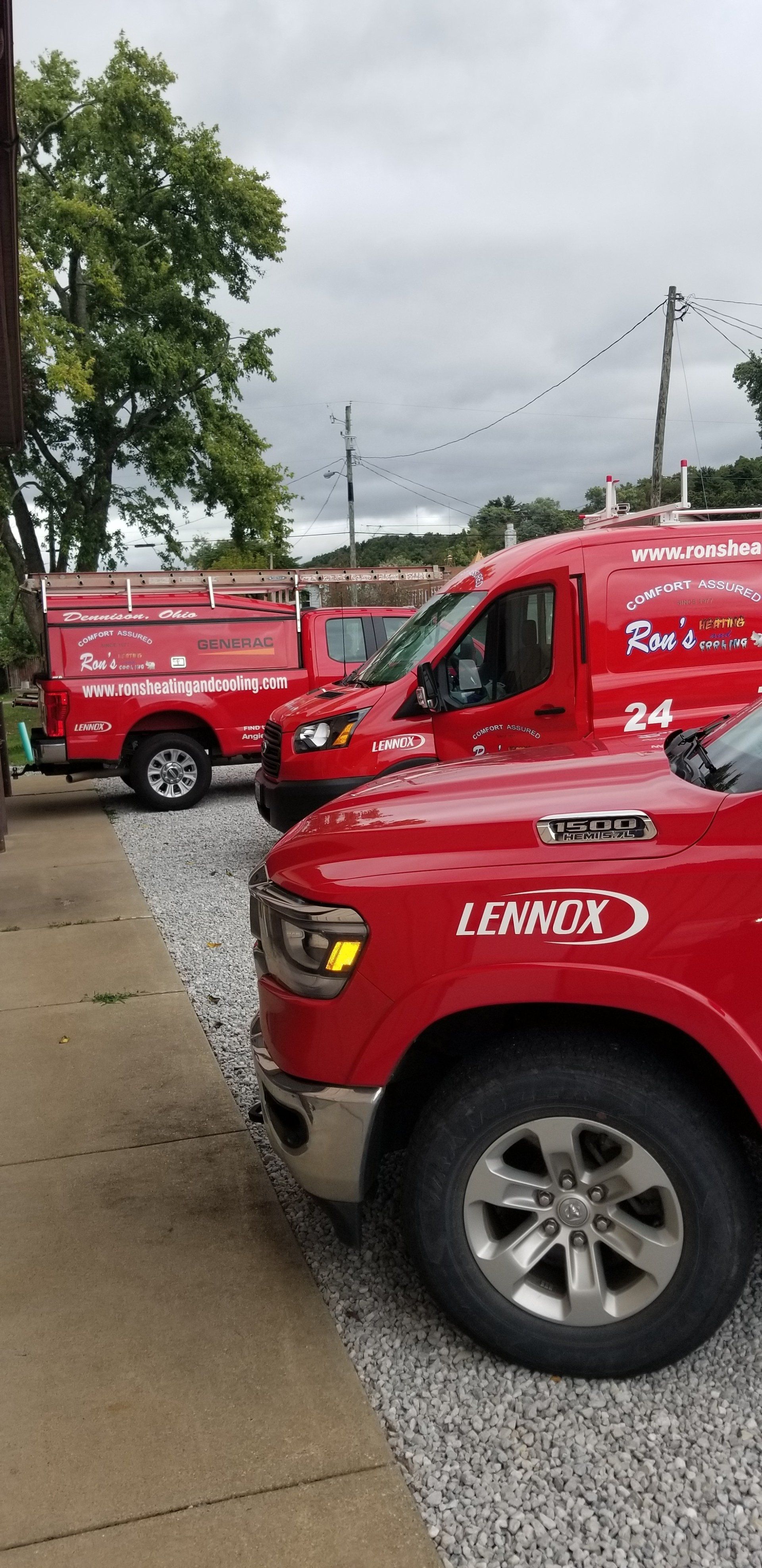 Red service vehicles parked on gravel and concrete near a building and foliage, under a cloudy sky.