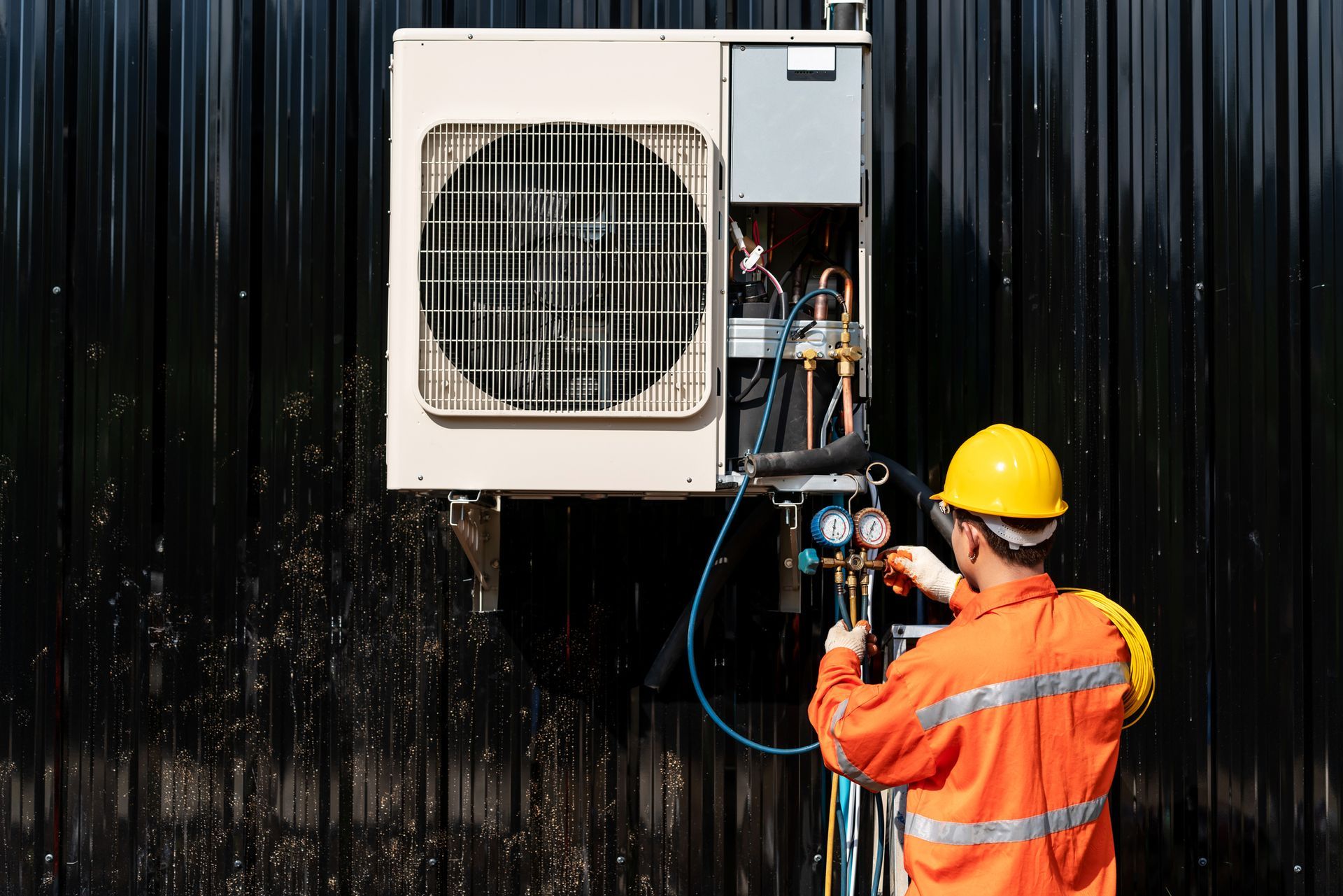 HVAC technician in orange uniform and yellow helmet repairs an air conditioning unit on a dark wall.