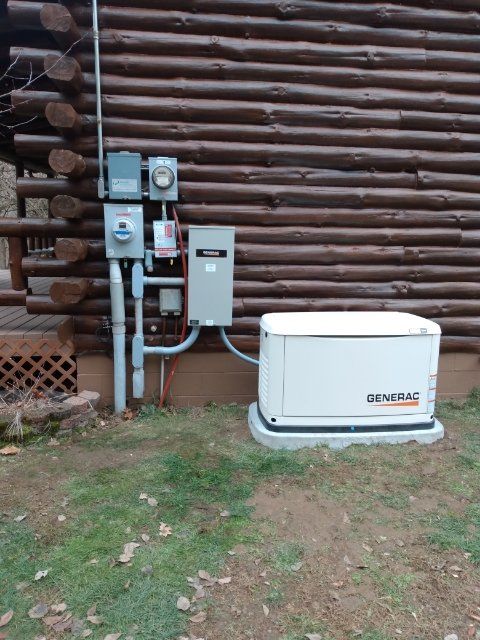 Generator and electrical components mounted on a log cabin exterior. Generator is white and gray, the other components are gray.
