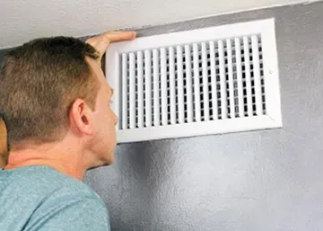 Cooling System and Duct Cleaning — Dennison, OH — Ron’s Heating & Cooling Man looking at a white air vent on a gray wall.