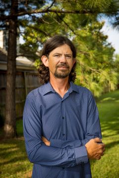 Man in a blue patterned shirt standing outdoors with arms crossed, in front of trees and a building.