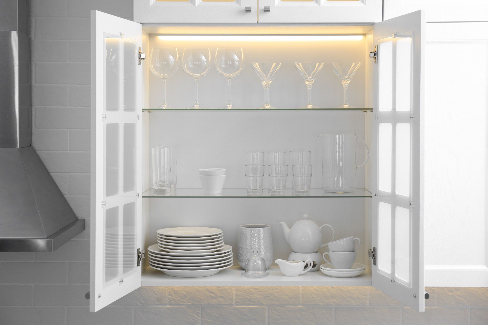 Open kitchen cabinet with glassware, plates, and bowls on white shelves, lit by warm under-cabinet lights