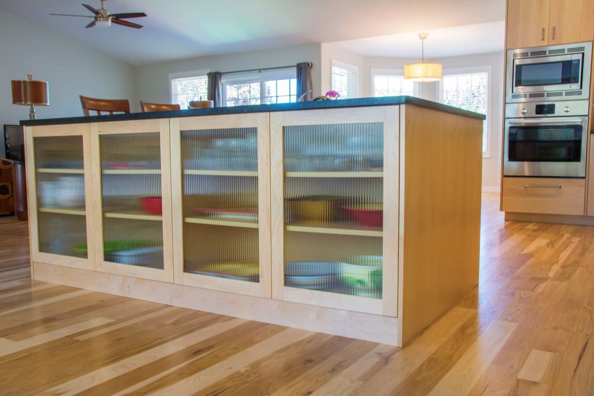 Modern kitchen with a large island featuring glass-front cabinets and built-in ovens in the background