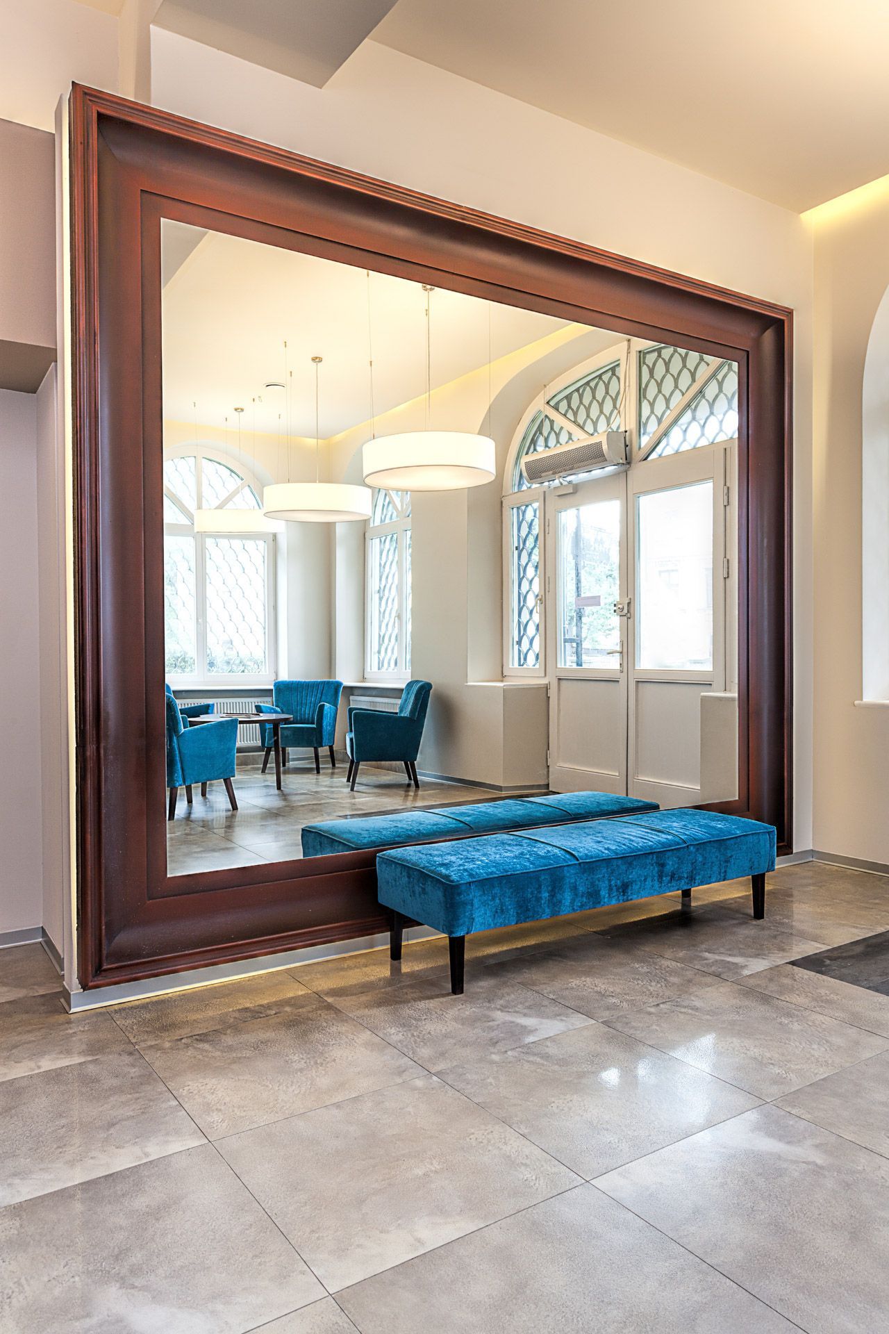 Modern lobby with a large framed mirror, blue bench, and blue chairs by tall arched windows.