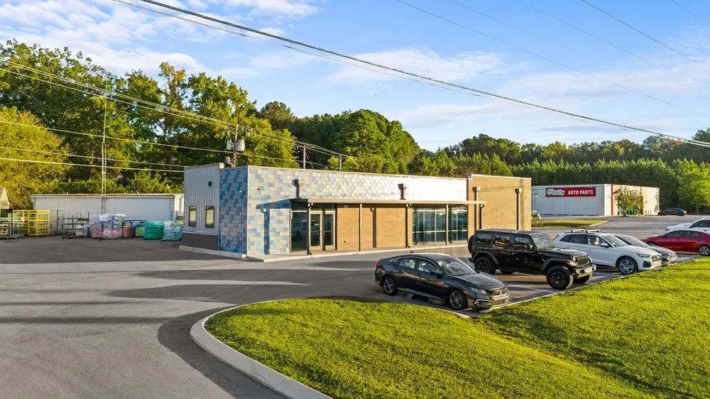 Small roadside business with parked cars, a gray building, and a grassy foreground under a blue sky